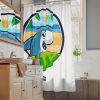 Kids bathroom shower curtain featuring a colorful cartoon shark with an ocean, beach, and sun design, shown in a bright modern bathroom with a vanity, mirror, and window.