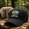 Black hiking dad hat with “Hike More Worry Less” mountain graphic placed on a tree stump outdoors with a metal camping mug in a blurred forest background.