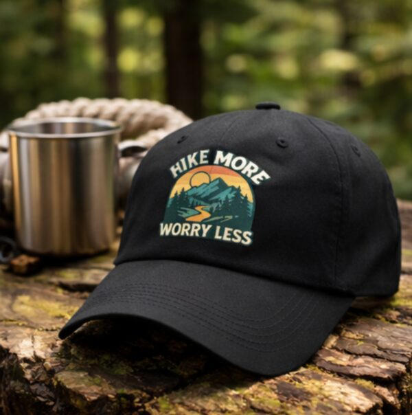 Black hiking dad hat with “Hike More Worry Less” mountain graphic placed on a tree stump outdoors with a metal camping mug in a blurred forest background.