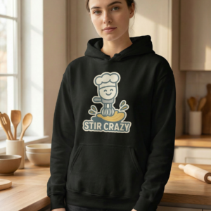theelegant-store-stir-crazy-funny-baking-hoodie-black-womens-kitchen Woman wearing a black “Stir Crazy” baking hoodie with a cute chef graphic, photographed in a warm home kitchen with wooden utensils and baking tools in the background.
