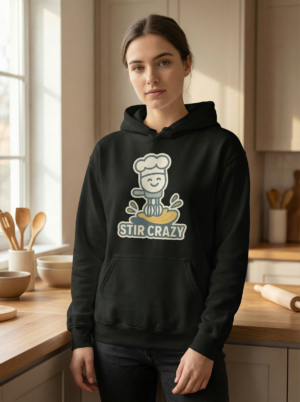 Woman wearing a black “Stir Crazy” baking hoodie with a cute chef graphic, photographed in a warm home kitchen with wooden utensils and baking tools in the background.