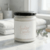 Scented candle in a clear glass jar with a black lid and pink script label on a marble counter beside towels and eucalyptus in a bright bathroom.