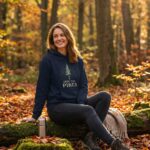 Male model wearing the Into the Pines unisex hoodie in navy with minimal pine tree graphic, standing in a sunlit forest.