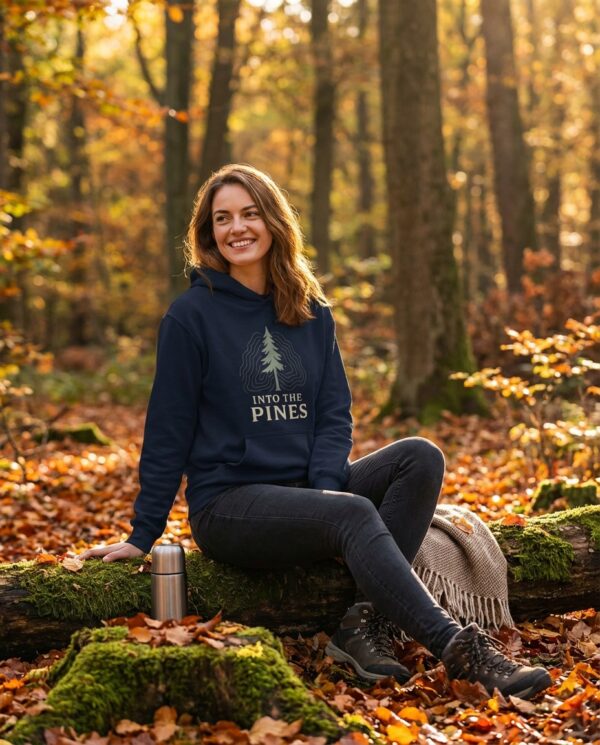 Male model wearing the Into the Pines unisex hoodie in navy with minimal pine tree graphic, standing in a sunlit forest.