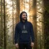 Male model in a sunlit forest wearing the Into the Pines unisex hoodie in navy featuring a minimal pine tree nature design.