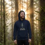 Male model in a sunlit forest wearing the Into the Pines unisex hoodie in navy featuring a minimal pine tree nature design.