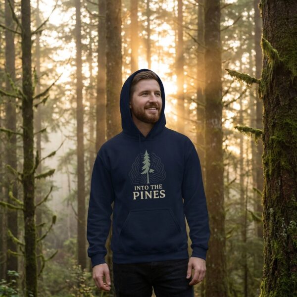Male model in a sunlit forest wearing the Into the Pines unisex hoodie in navy featuring a minimal pine tree nature design.